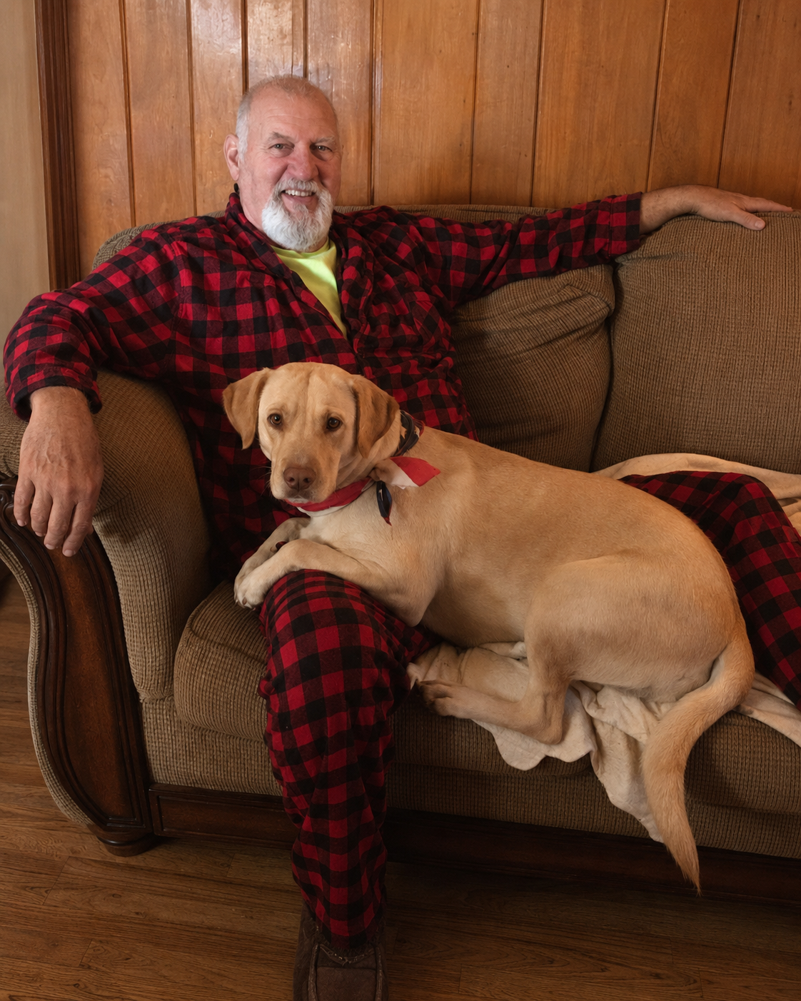 Older man in red plaid loungewear smiling on a couch while a yellow Labrador rests beside him.